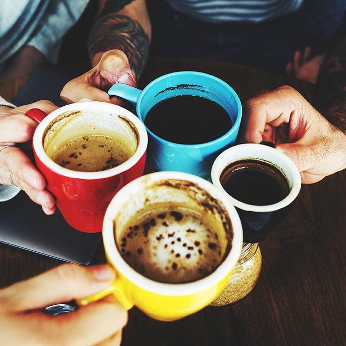 Close-up of hands holding colorful coffee mugs filled with various types of coffee, raised in a toast. The setting conveys warmth, camaraderie, and a cozy gathering.