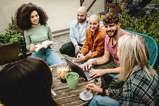 Group of young adults sitting around a table outdoors, engaged in conversation and collaboration. One person is using a laptop, while others are smiling, holding books, or sipping coffee. The setting includes greenery, creating a relaxed and inviting atmosphere.